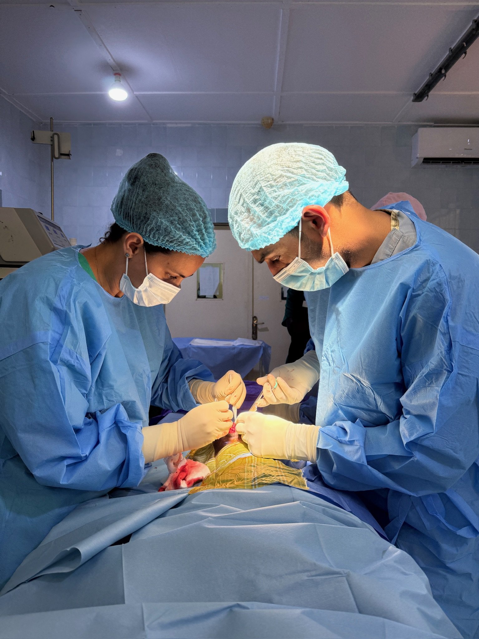 Surgeons performing an operation in a hospital operating room, wearing surgical scrubs, masks, and gloves, with medical equipment in the background.