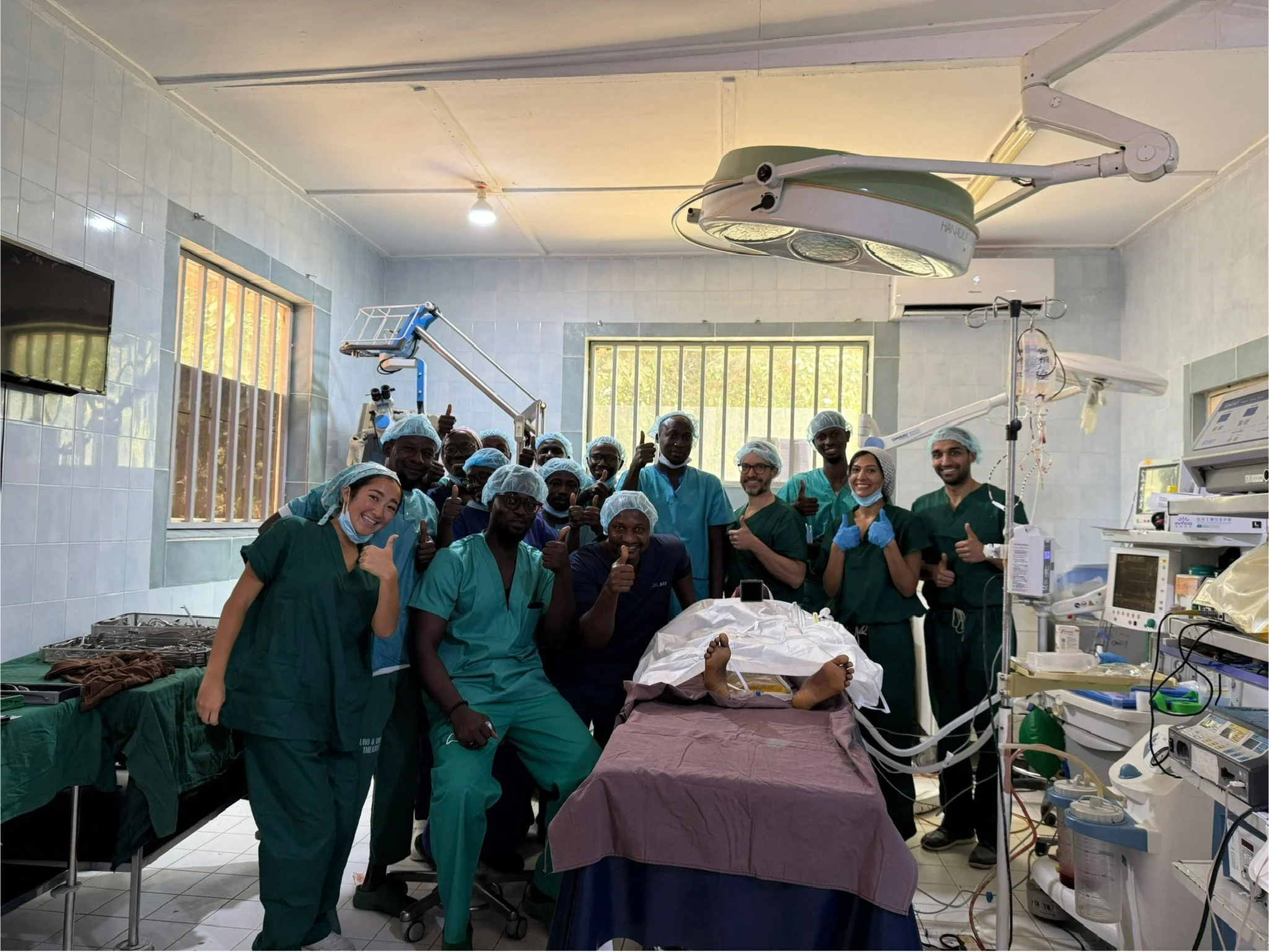 Group of medical professionals and staff in a hospital operating room gathered around a patient on an operating table, some giving thumbs up, with medical equipment and monitors around.