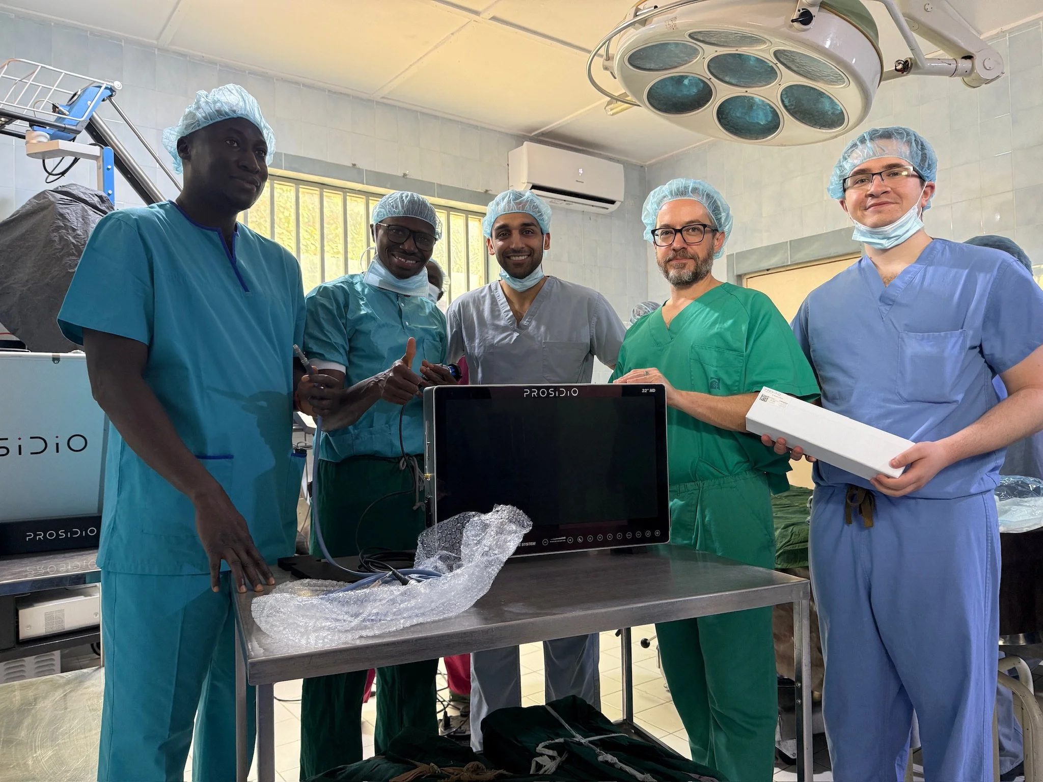Group of five medical professionals in scrubs and surgical caps standing in an operating room, gathered around medical equipment and a computer monitor.