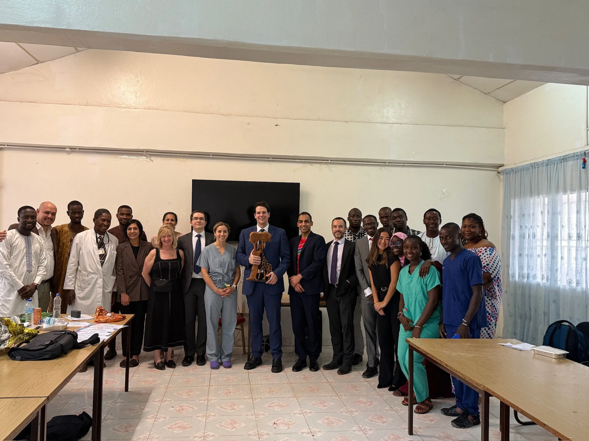 Group of diverse professionals and students standing together in a classroom or conference room, smiling at the camera, with tables and a monitor in the background.