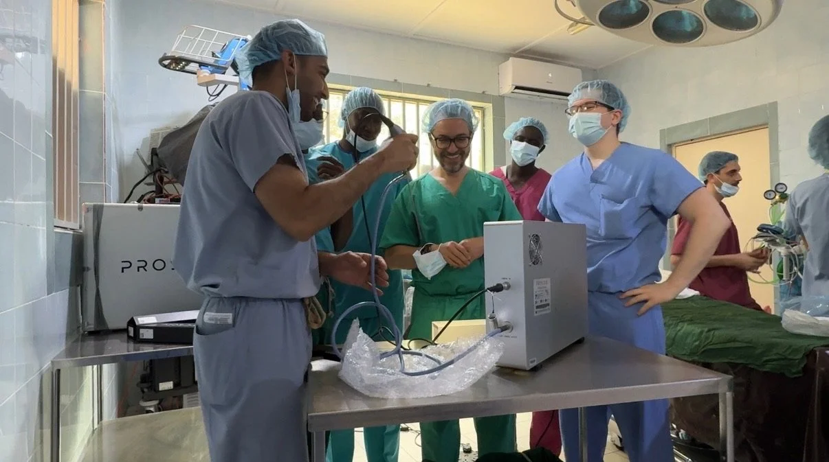 Group of medical professionals in scrubs and masks gathered around medical equipment in an operating room, smiling and conversing.