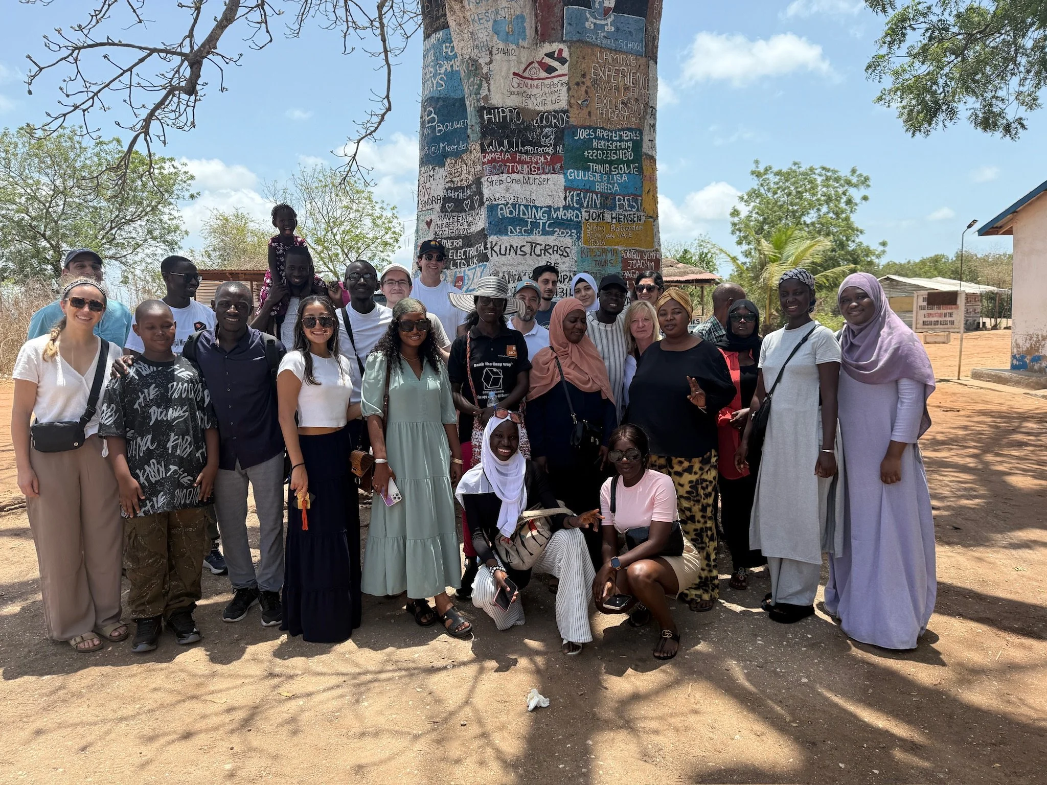 A diverse group of people, including men, women, and children, gathered outdoors in front of a large painted tree trunk with colorful messages. The group appears happy, smiling, and dressed casually under a bright blue sky with some trees and buildin