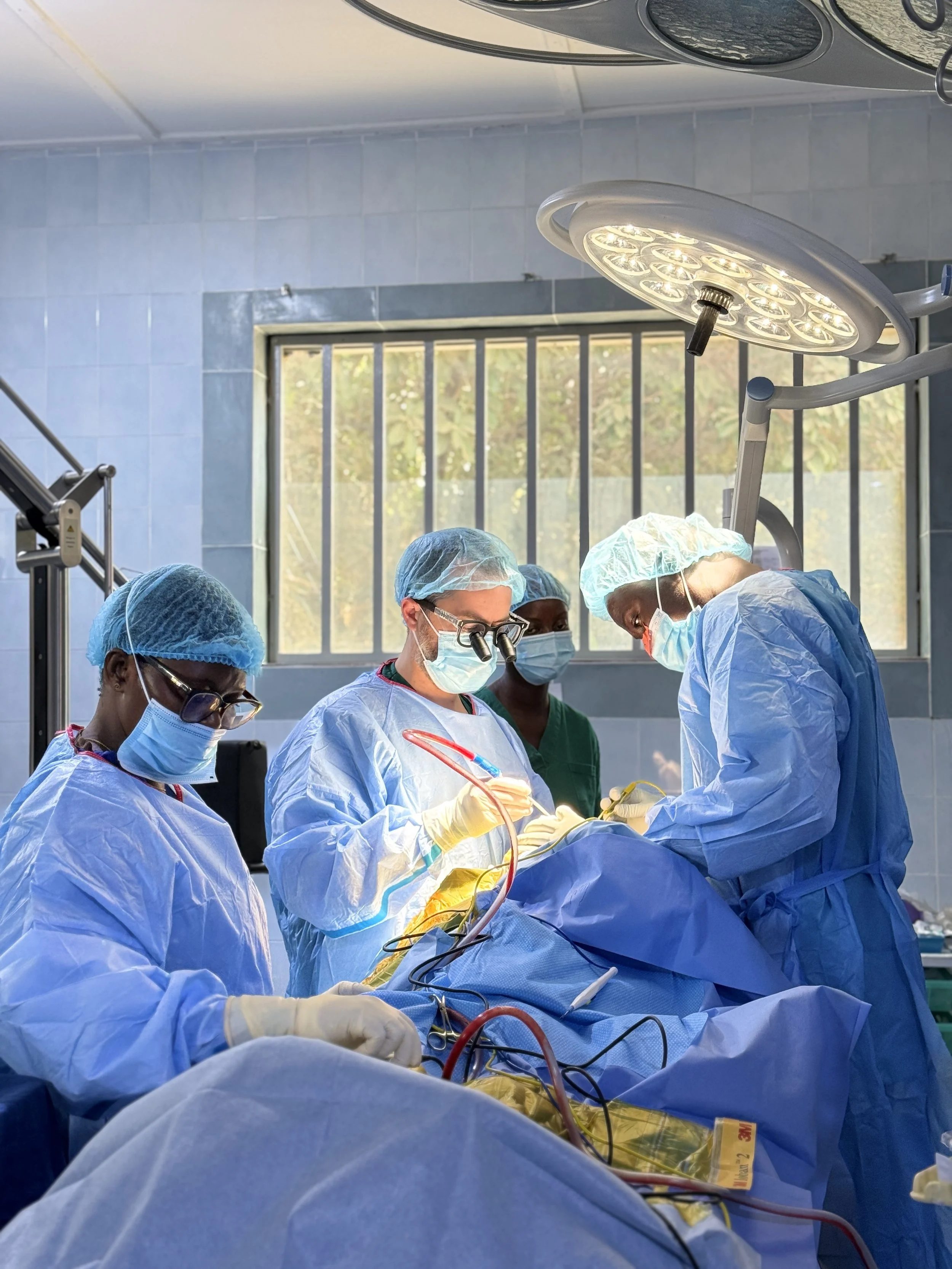 Medical team performing surgery in an operating room with overhead surgical light and window in background.