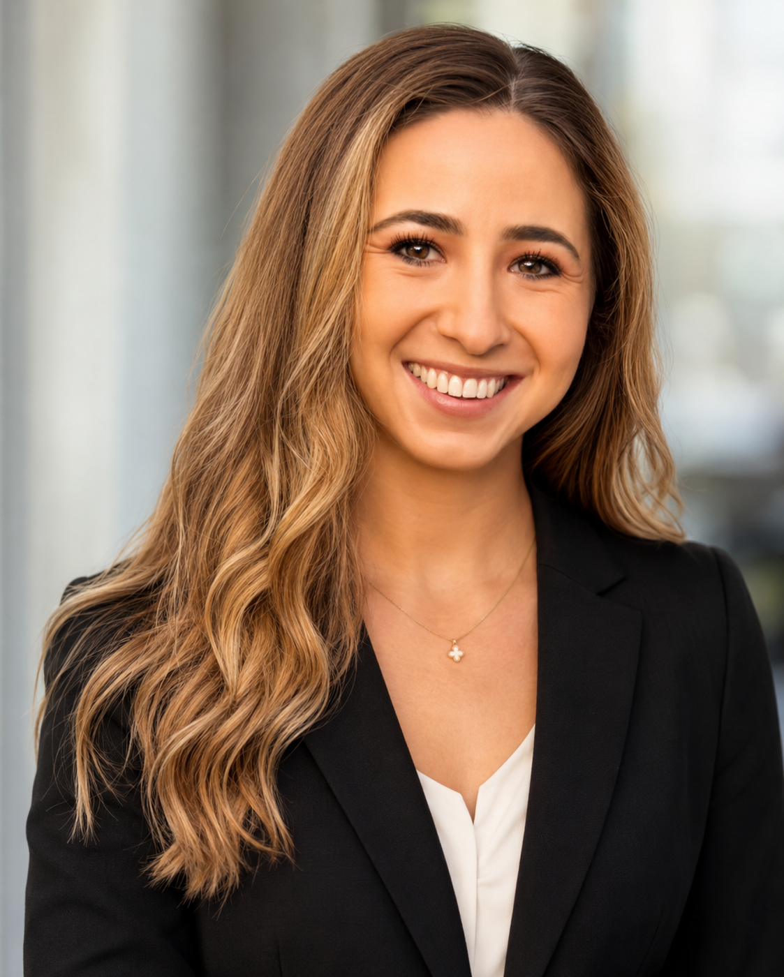 Professional woman with long wavy hair, smiling, wearing a black blazer, white blouse, and a gold necklace with a four-leaf clover pendant, standing in an office setting.