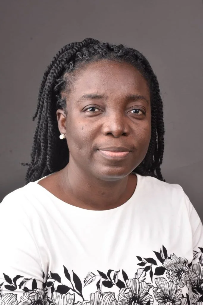 Portrait of a woman with dark skin and braided hair, wearing a white shirt with black floral designs and pearl earrings, against a plain dark grey background.