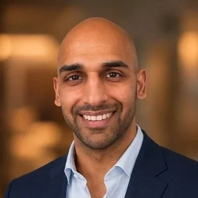 A smiling man with a shaved head, wearing a navy blazer and light blue shirt, in a professional setting