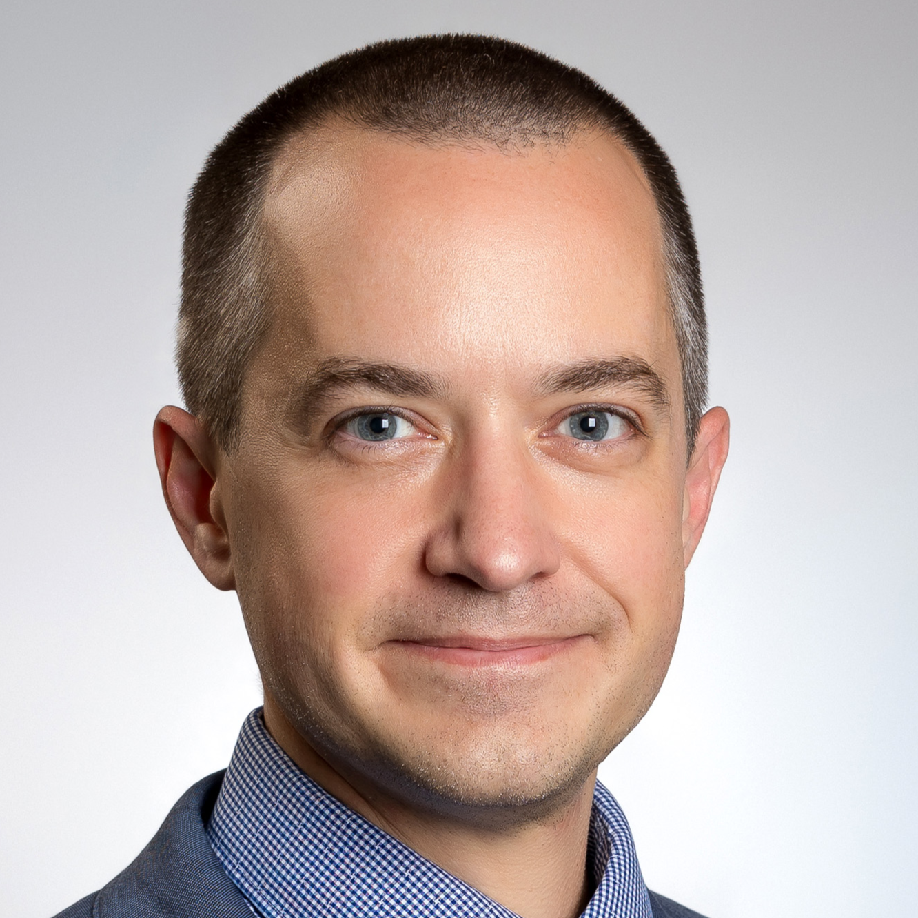 A professional headshot of a man with short dark hair and blue eyes, wearing a blue checkered shirt and a dark blazer, smiling slightly against a plain light gray background.