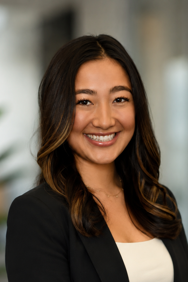 Close-up portrait of a young woman with long dark hair, smiling, wearing a black blazer and a white top, with a blurred office background.