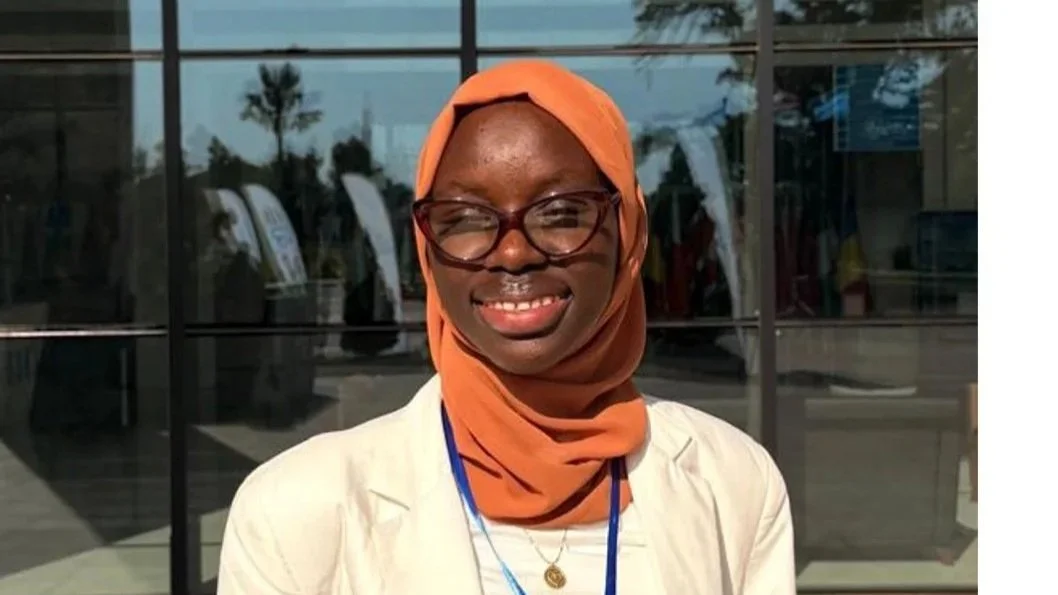 A smiling woman with glasses and a orange headscarf standing outside a glass building.