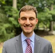 A young man in a gray suit and pink striped tie smiling outdoors with green trees in the background.