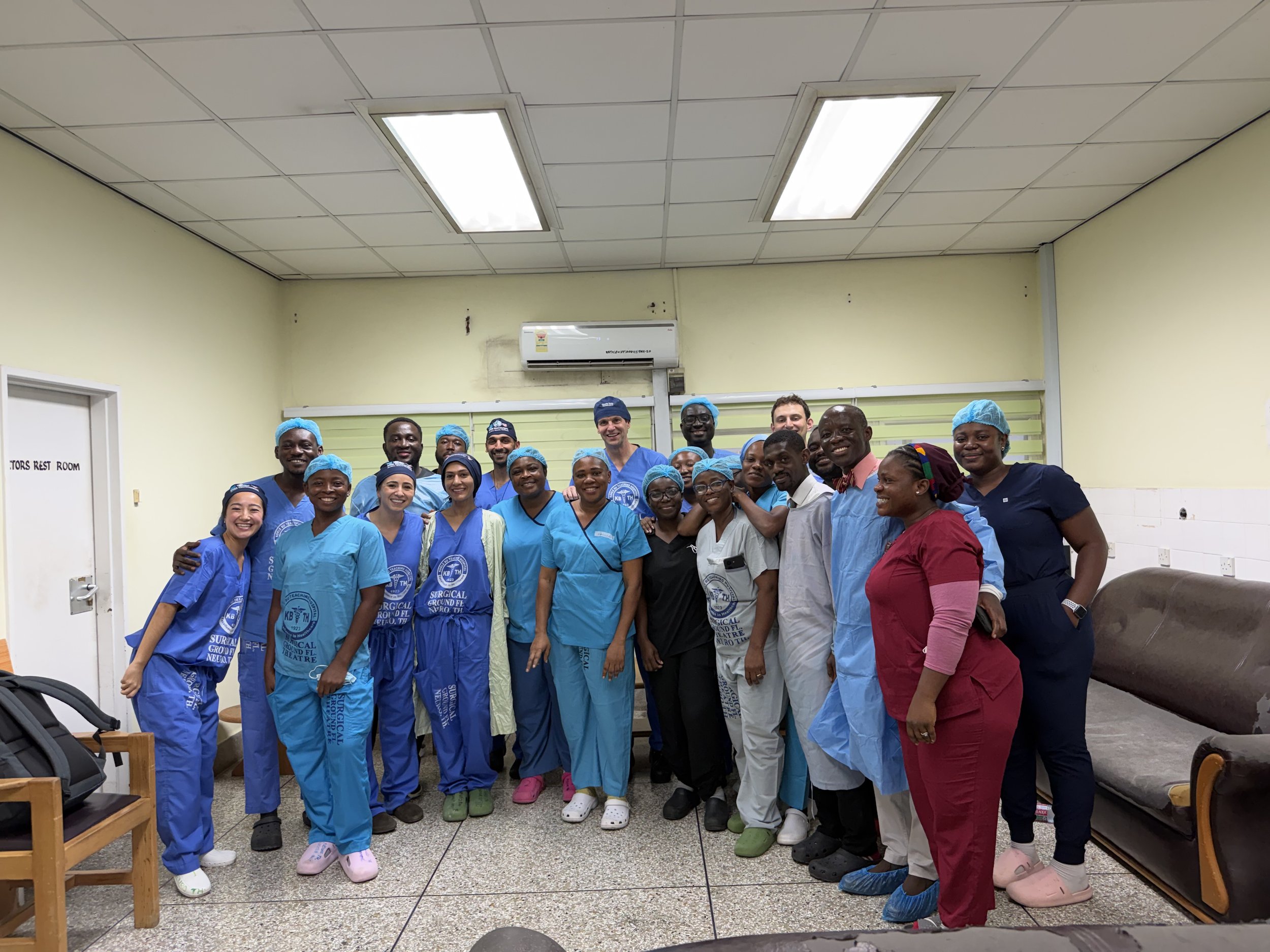 Group of healthcare professionals and staff posing together in a hospital or clinic room, wearing scrubs and hair covers, smiling for the photo.
