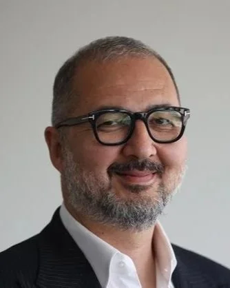 A middle-aged man with glasses, a short beard, and a slight smile, dressed in a dark suit jacket and white shirt, in front of a plain light gray background.