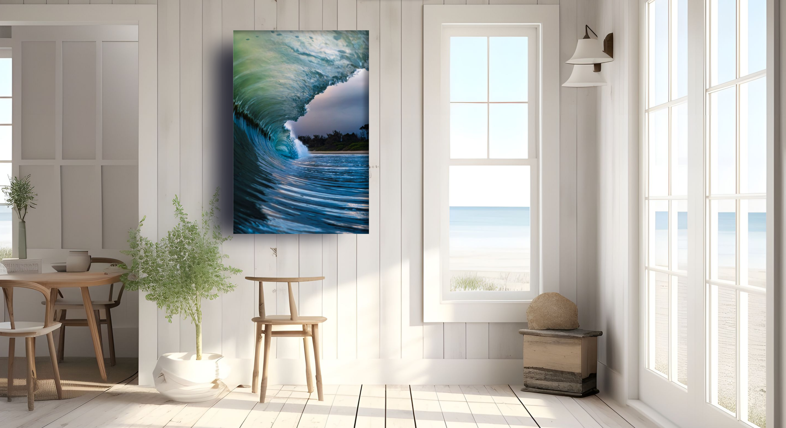 Interior of a bright room with white wooden walls and large windows showing a view of the beach and ocean. A large painting of a wave hangs on the wall. There are some potted plants and a small wooden table with chairs.