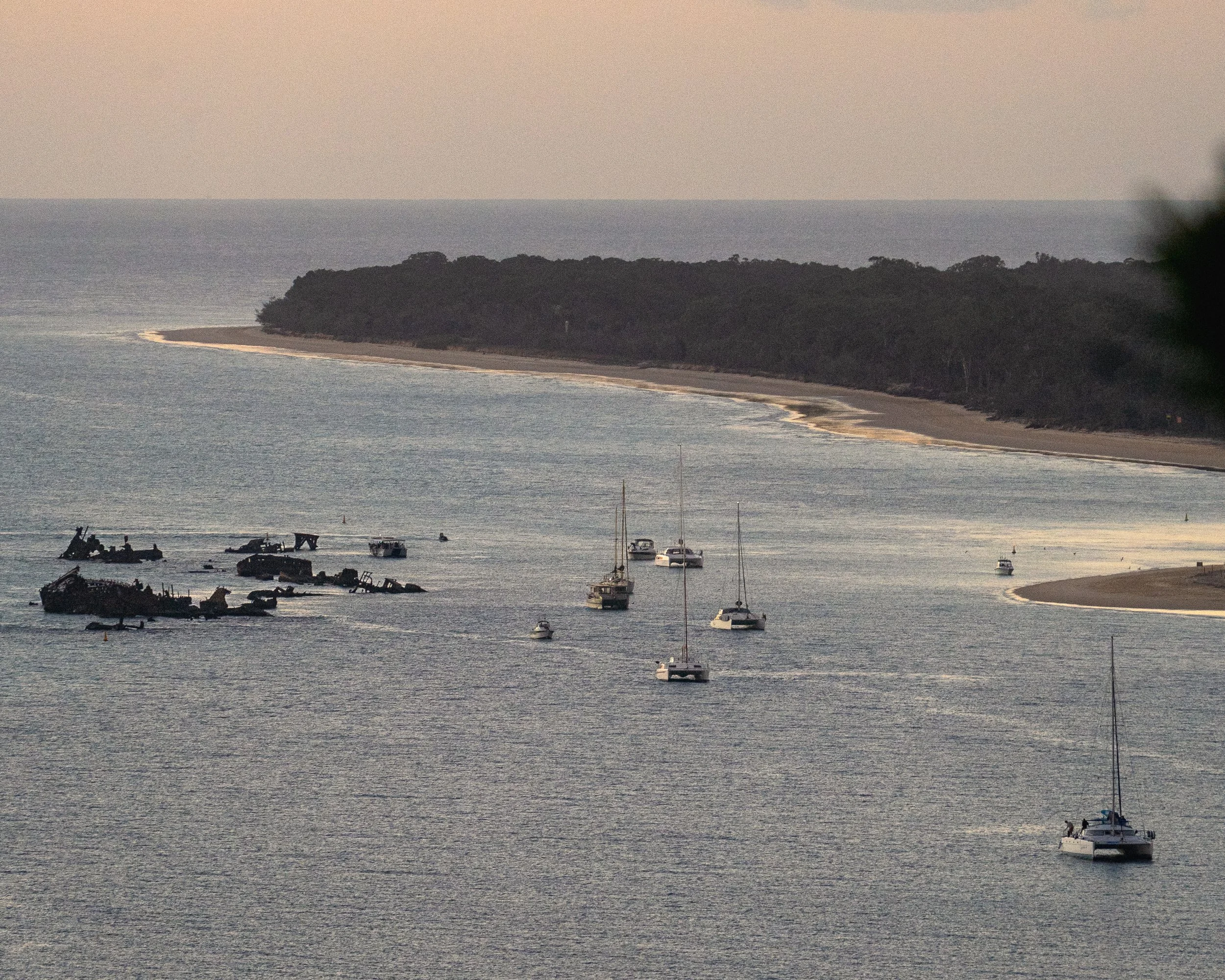 A body of water with several boats anchored near the shore, and a wooded coastline in the background during dusk or dawn.