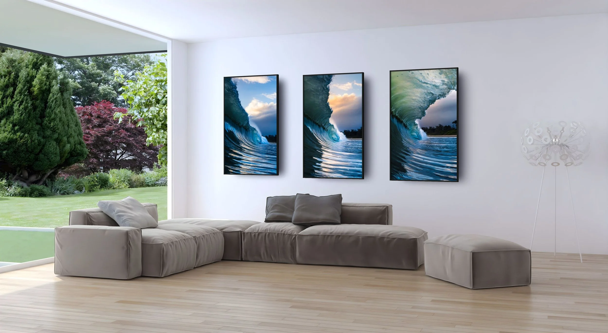 Modern living room with large windows overlooking a garden, a beige sofa with pillows, and three wall-mounted digital art displays of ocean waves.
