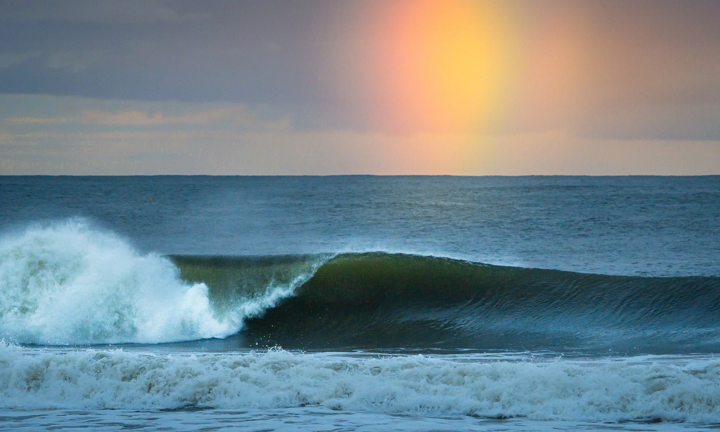 Ocean waves with a colorful rainbow appearing in the sky above the horizon at sunset or sunrise.