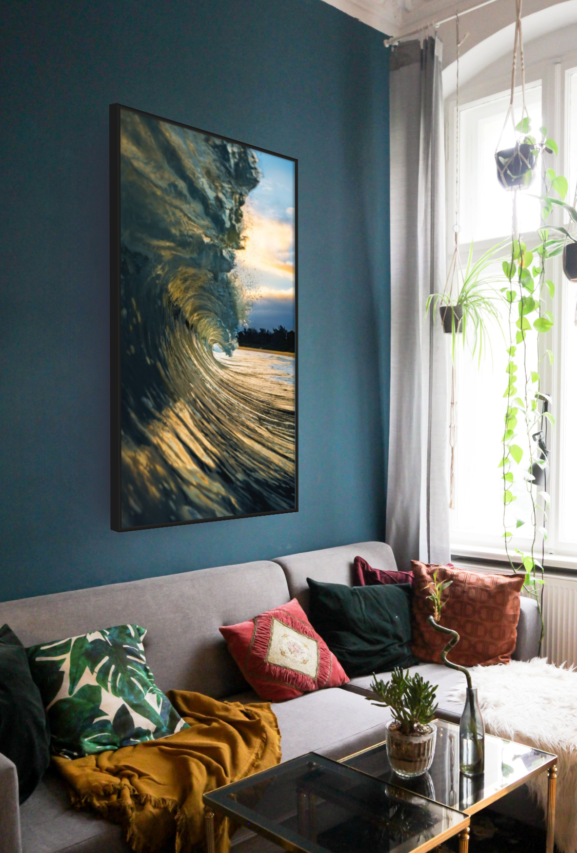Living room with a blue accent wall, a borderless flat-screen TV displaying an ocean wave at sunset, a gray sofa with multicolored pillows, a glass coffee table with a small plant, and hanging potted plants near a large window with white curtains.