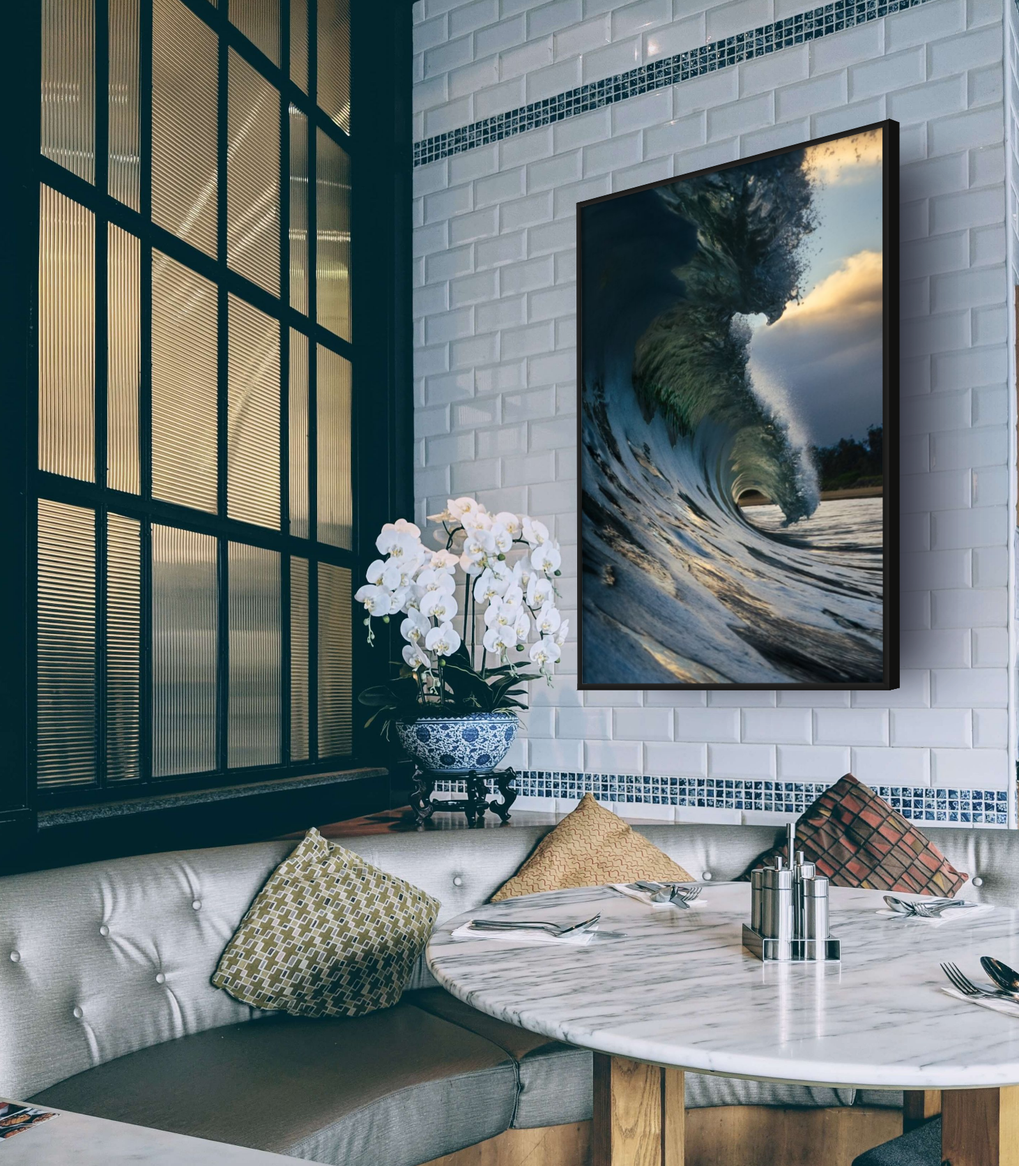 Interior view of a dining area with a marble-top round table, patterned and solid cushions on a sofa, a framed photograph of a large wave and tree on the wall, a potted white orchid plant, and a window with black grid frames and textured glass.
