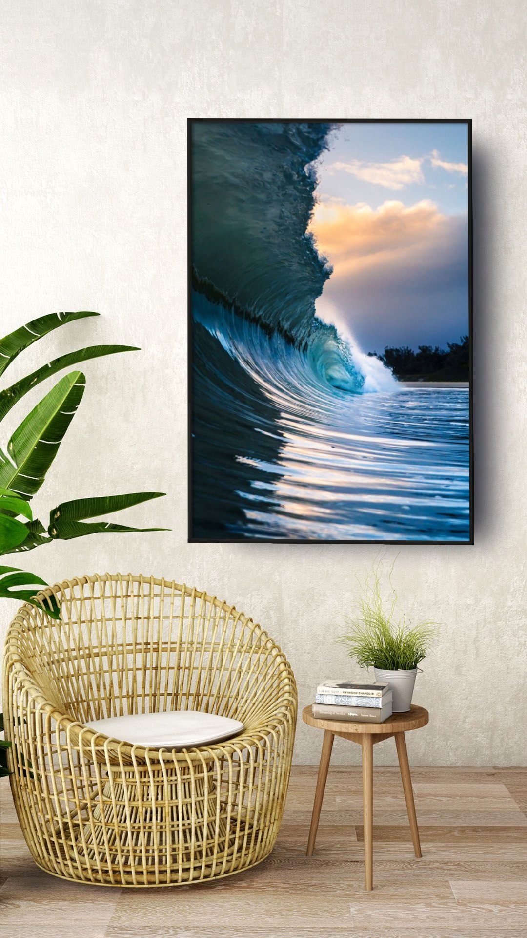 A framed photo of an ocean wave at sunset hangs on a light-colored textured wall, with a green plant, wicker chair with a cushion, and a small wooden table holding a stack of books and a potted plant in a home interior.