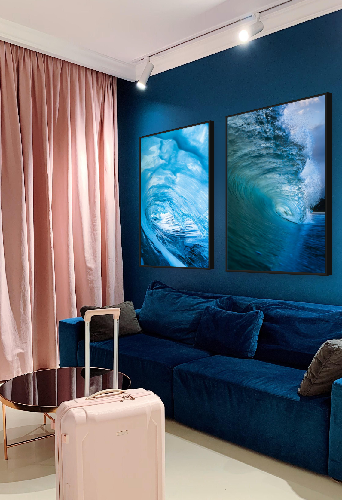 A hotel room with pink curtains, a blue velvet sofa, and two framed ocean wave pictures on a teal wall. There is a pink suitcase and a round glass table in front.