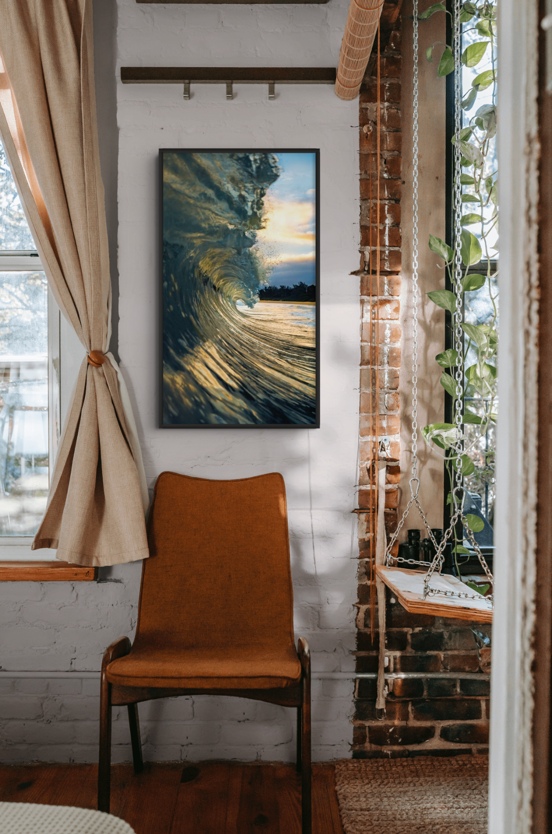Interior of a cozy room featuring a framed ocean wave photograph on a white brick wall, a brown mid-century armchair, a window with beige curtains, and a hanging wooden swing chain with green plants beside a brick column.