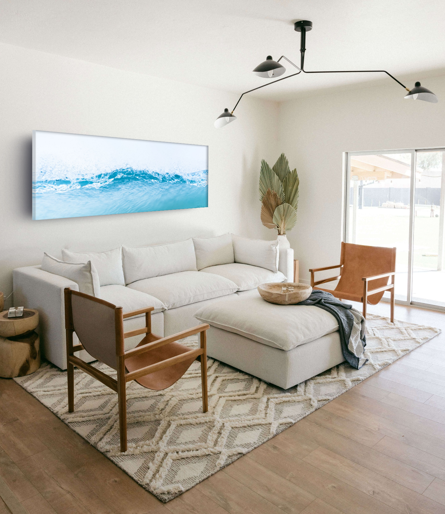 Living room with white sofa, wooden chairs, a patterned rug, large wall art depicting waves, a potted plant, a round wooden coffee table, and sliding glass door letting in natural light.
