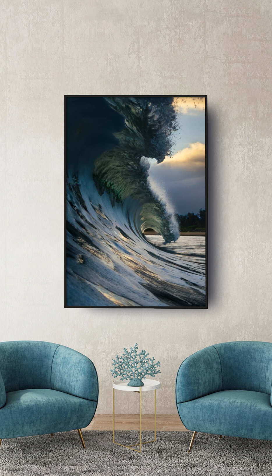 A framed photo of an ocean wave crashing, with a cloudy sky in the background, hanging on a beige wall in a living room with two teal chairs and a small white side table with a decorative teal coral piece.