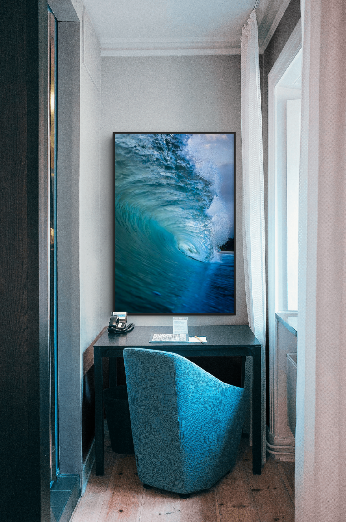 Small hotel room corner with a desk, a blue patterned armchair, a large wall-mounted television displaying a wave, a telephone, a notepad, and a pen on the desk, with a window and white curtains on the right.