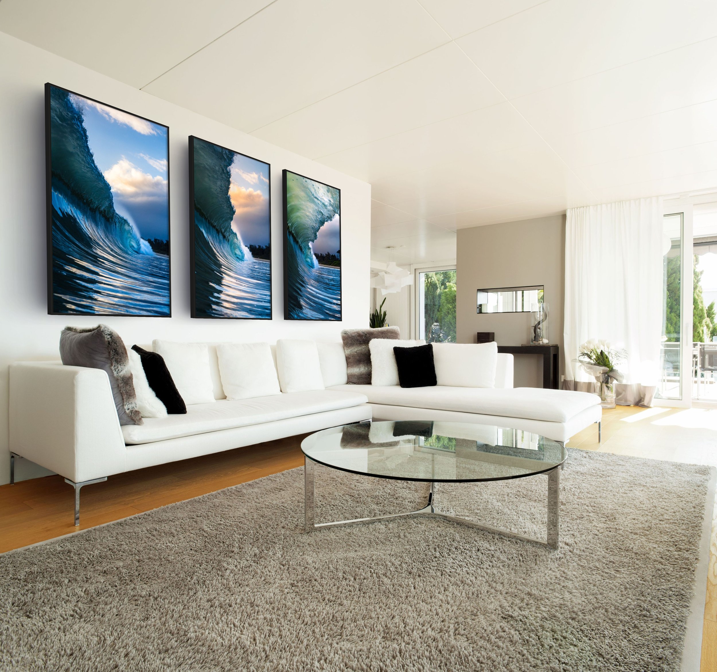 Living room with white sectional sofa, black and white pillows, round glass coffee table, shaggy beige carpet, large windows with white curtains, and three framed wave paintings on white wall.