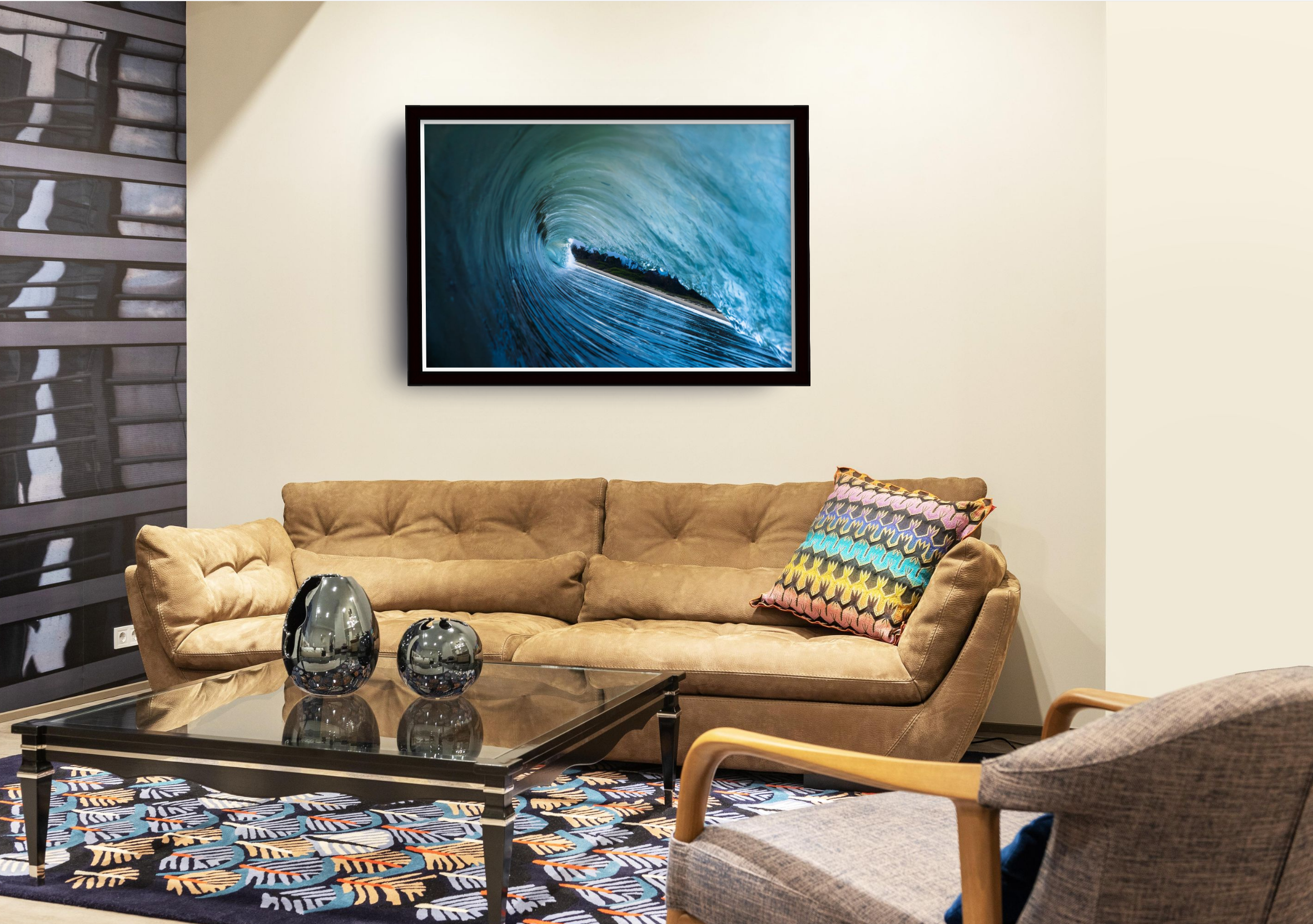 Living room with beige tufted sofa, colorful patterned pillow, black glass coffee table with three decorative vases, patterned rug, cream wall with a large framed ocean wave photograph, window with black sectional blinds to the left, wooden armchair at the front right corner.