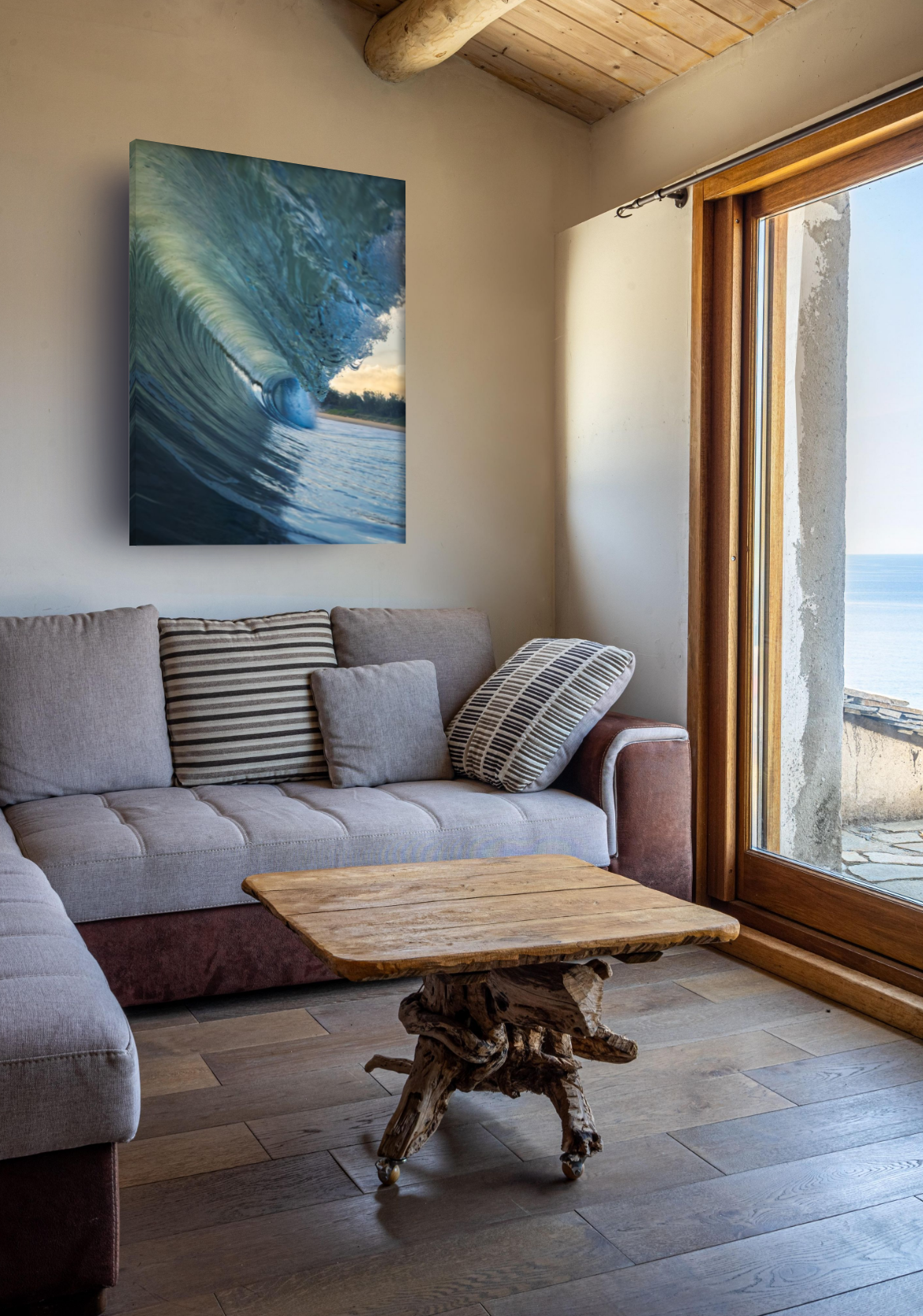 Living room with a beige sofa, striped and plain cushions, a rustic wooden coffee table, a large window showing a view of the ocean, and a wall hanging of a large wave.