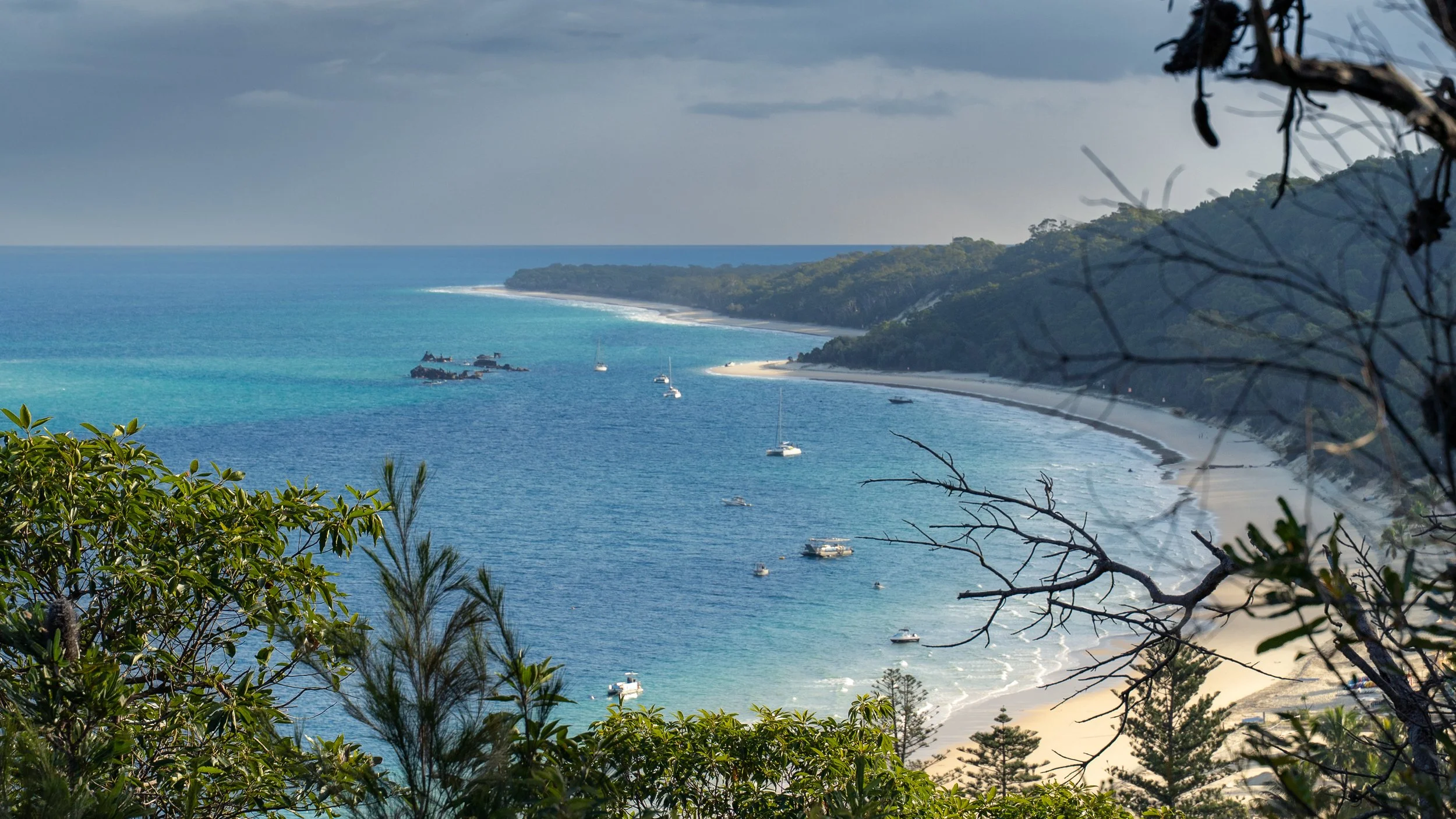 A scenic view of Tangalooma on Queensland's Moreton Island capturing the wrecks and shoreline, surrounded by lush green trees and hills under a cloudy sky.