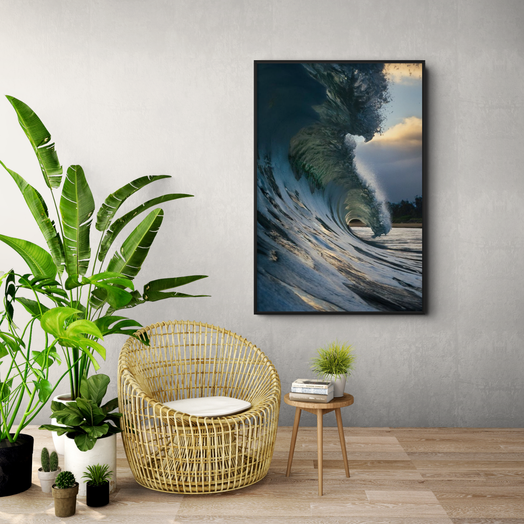 Living room with a large framed ocean wave photograph on a light gray wall, a wicker chair with a white cushion, a small wooden side table with a stack of books and a small potted plant, additional small potted plants on the floor, and a large green houseplant to the left.