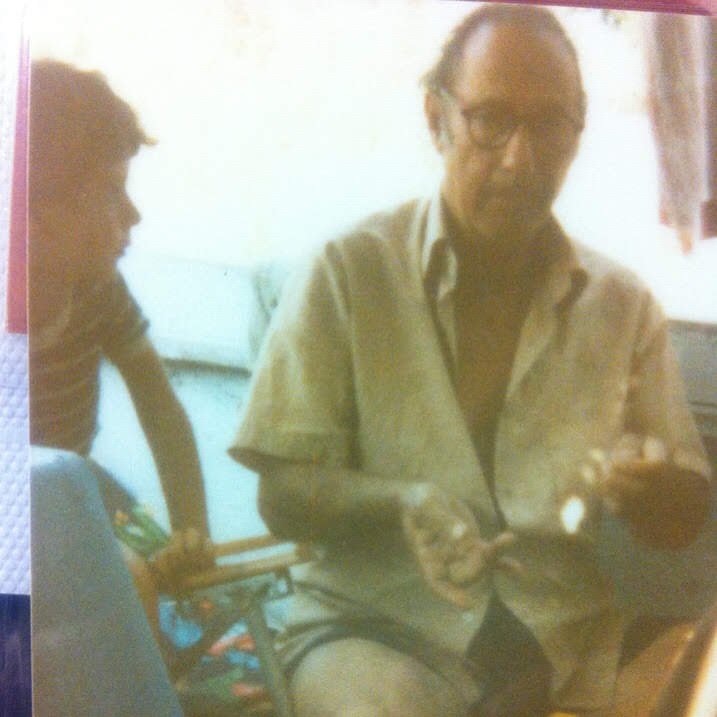 Two men sitting indoors, one with glasses, wearing a beige shirt, and the other with dark hair, wearing a dark shirt, engaged in conversation.