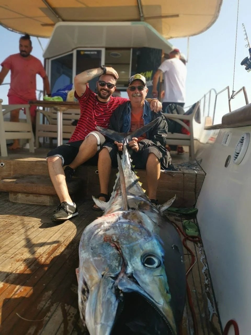 Two men sitting on the deck of a boat, smiling and holding a large marlin fish they caught. One man is wearing sunglasses and a baseball cap, the other is wearing a red and black striped shirt. In the background, two other people are near the boat's cabin.