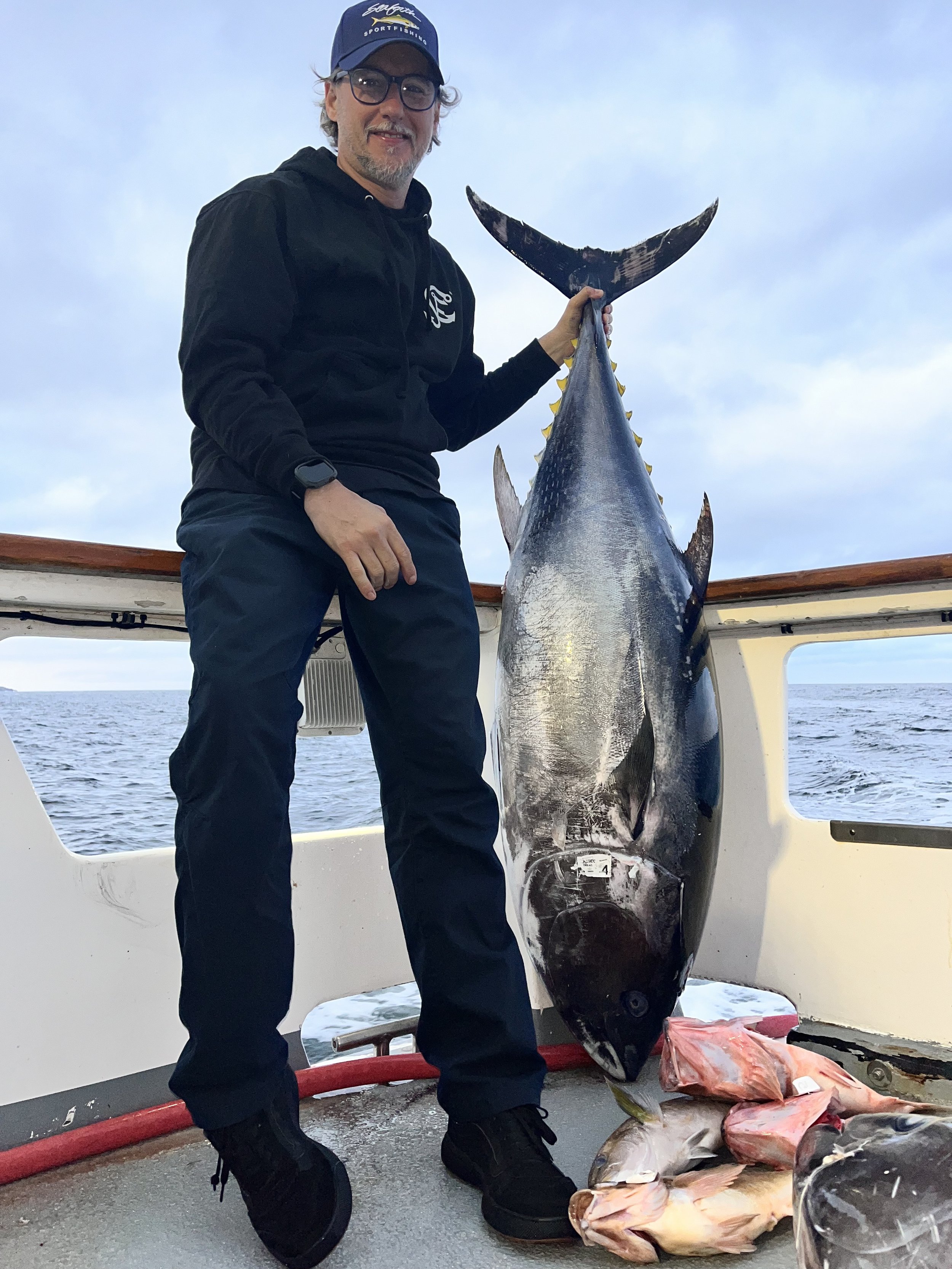 A man wearing a black hoodie, glasses, and a blue cap is holding a large fish on a boat. The fish is black with a silvery belly and yellow fins, and there are several smaller fish laid out on the deck in front of him. The background shows the open ocean and a partly cloudy sky.