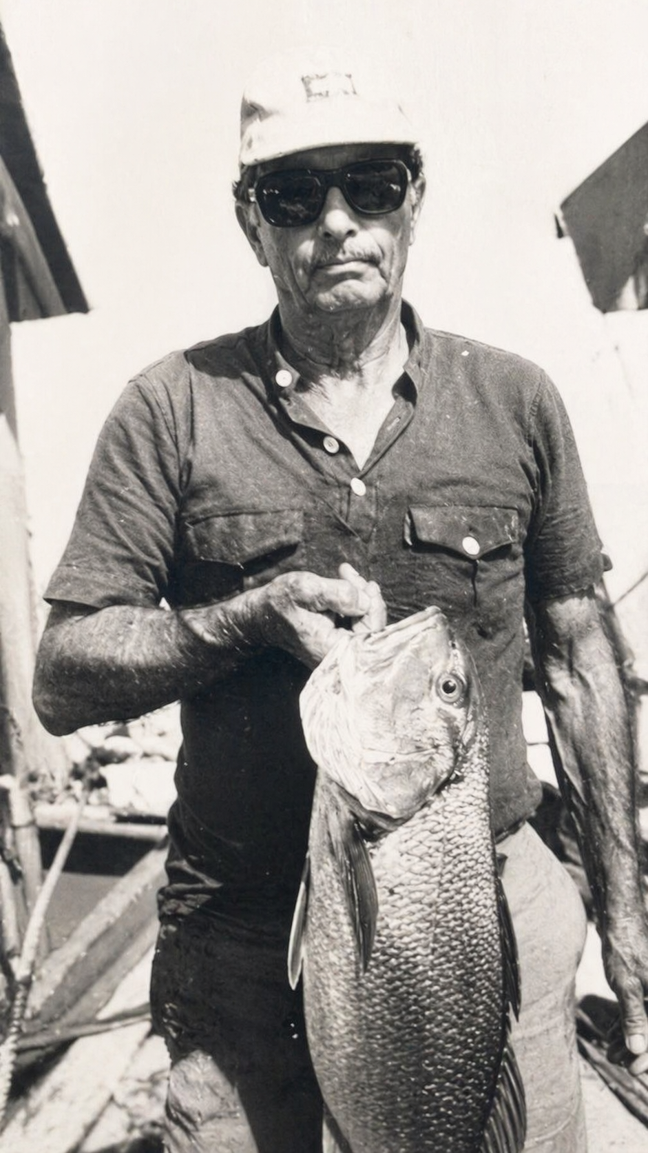 A man wearing sunglasses, a cap, and a short-sleeved shirt holds a large fish with his right hand.