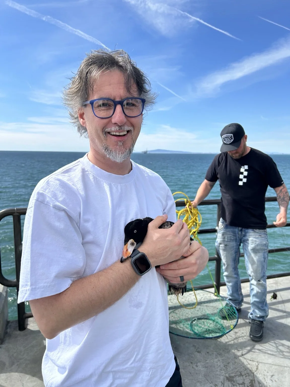 A smiling man with glasses holding a young duck on a boat against a backdrop of water and blue sky with clouds and contrails. A man in the background is handling yellow ropes.