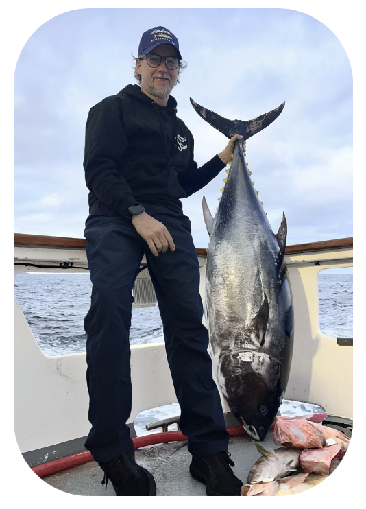 A man on a boat holding a large fish, with smaller fish on the deck, under a cloudy sky.