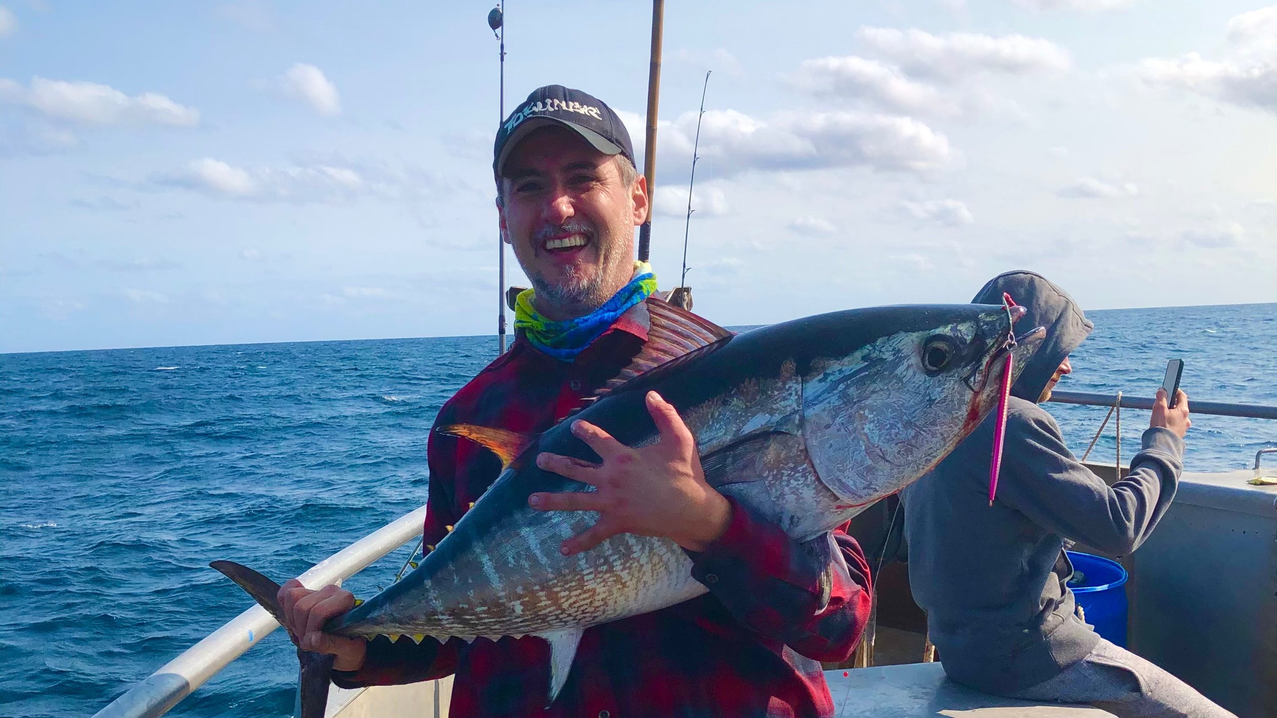 A man smiling and holding a large fish on a boat out at sea, with another person in the background using a phone.