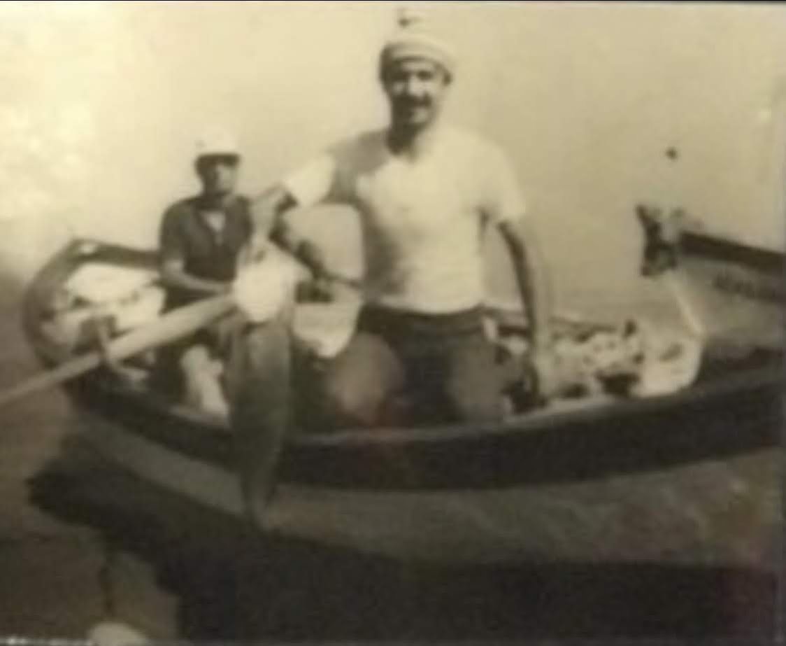 Black and white photograph of two men standing in a small boat, one man is holding an oar, and the boat appears to be on water with a plain background.