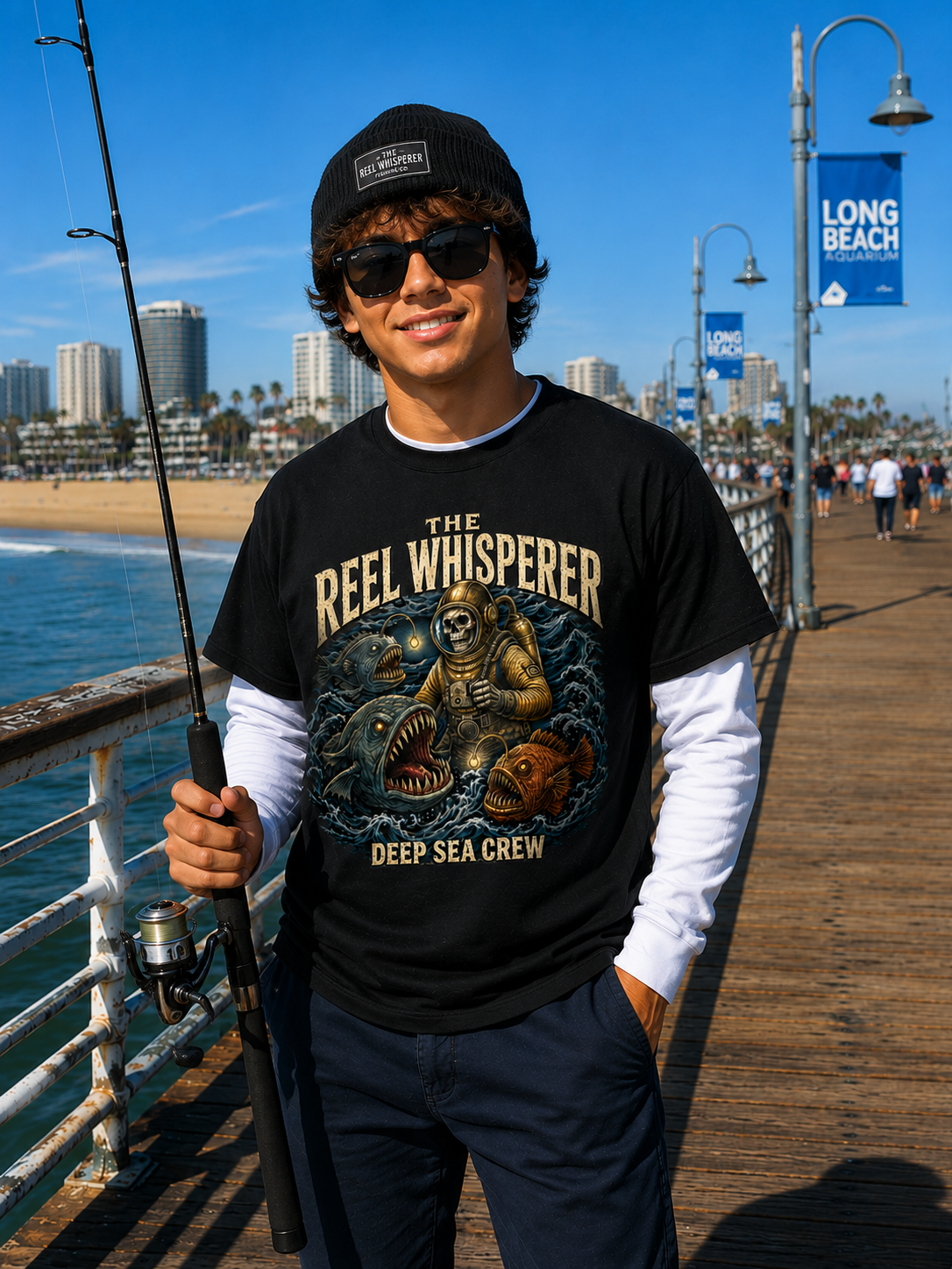 A young man on a pier holding a fishing rod, dressed in a black 'Reel Whisperer' t-shirt, black beanie, and sunglasses, with a seaside cityscape and blue sky in the background.