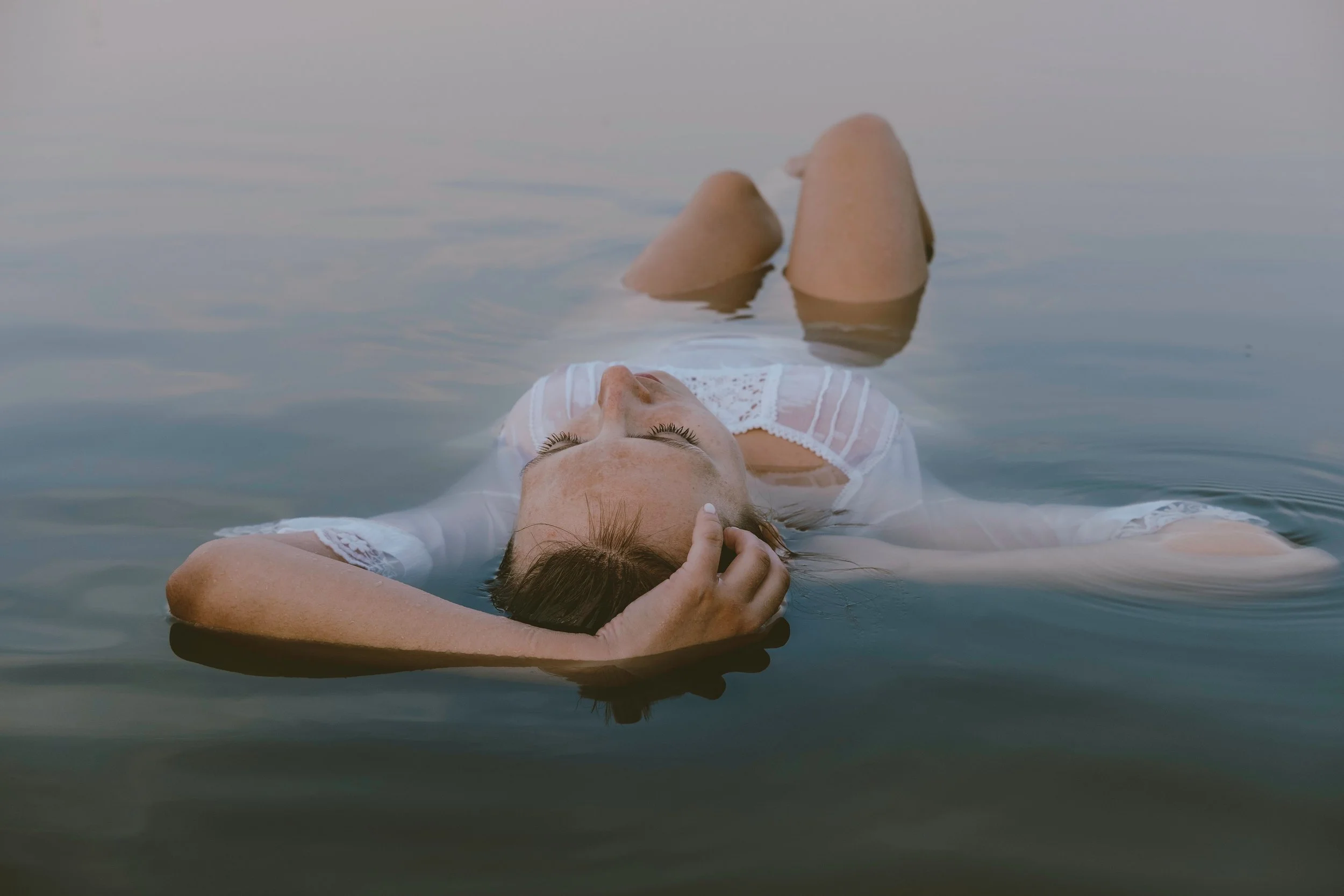A woman with brown hair and freckles lying on water, wearing a white lace top, with her eyes closed, one hand touching her forehead, immersed in water.