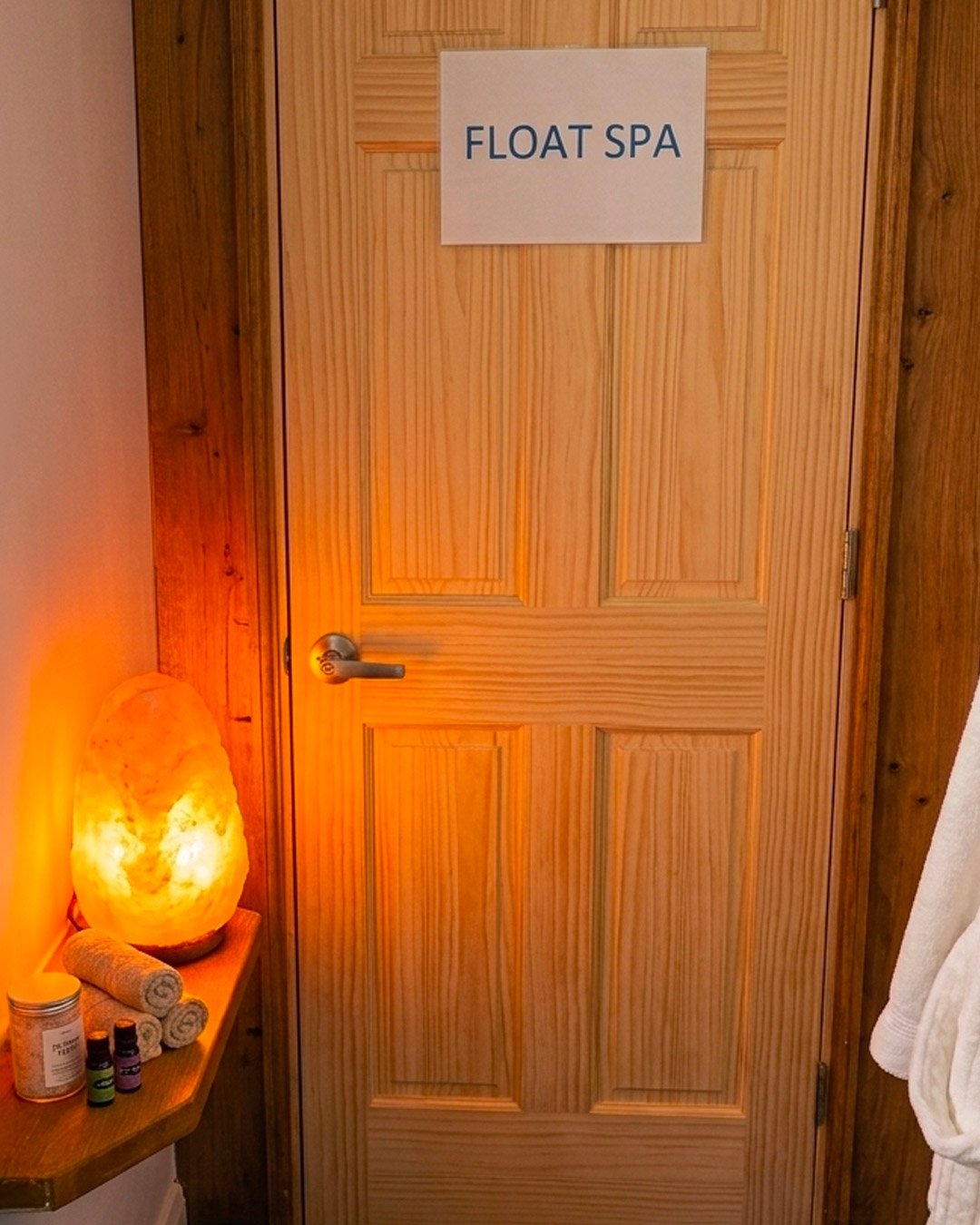 A wooden door with a sign that says 'Float Spa', lit by a salt lamp on a small table to the left of the door.