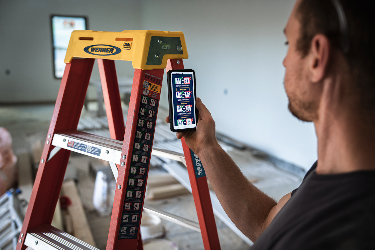 A man using a smartphone to scan a color-coded safety instruction sticker on a red and yellow Werner ladder in an indoor construction site.