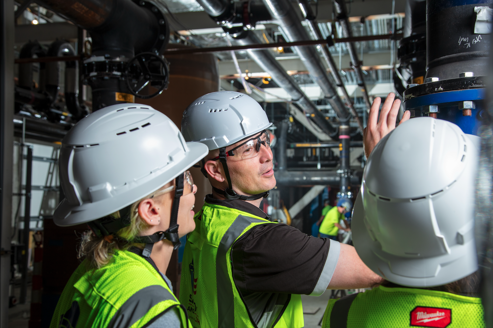 Group of construction workers in safety vests and helmets inspecting machinery indoors.