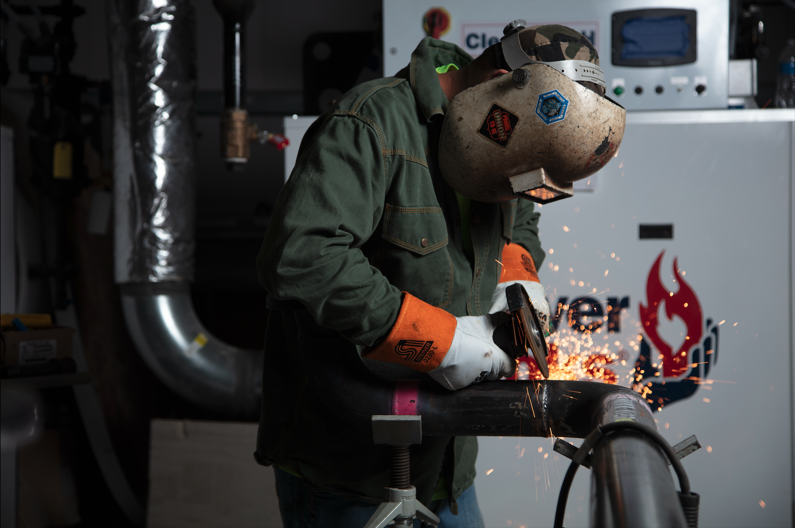 A worker welding metal pipe indoors, wearing protective gloves, a welding helmet, and a green work jacket, with sparks flying from the welding process.