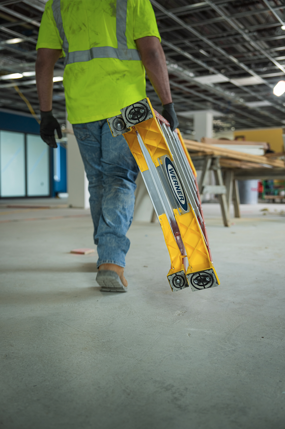 A construction worker walking inside a building under construction, carrying a yellow Werner safety tool with wheels, wearing a bright yellow safety vest, jeans, and work boots.