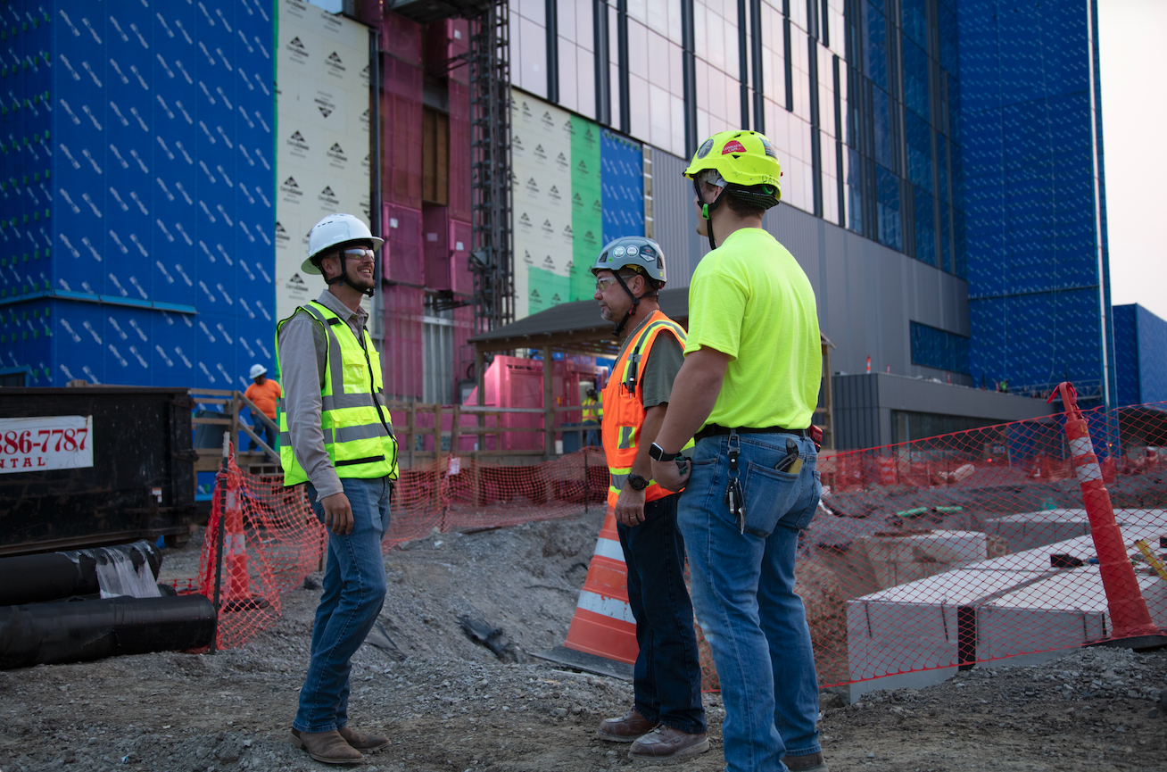 Three construction workers wearing safety helmets and vests are having a conversation on a construction site in front of a modern building.