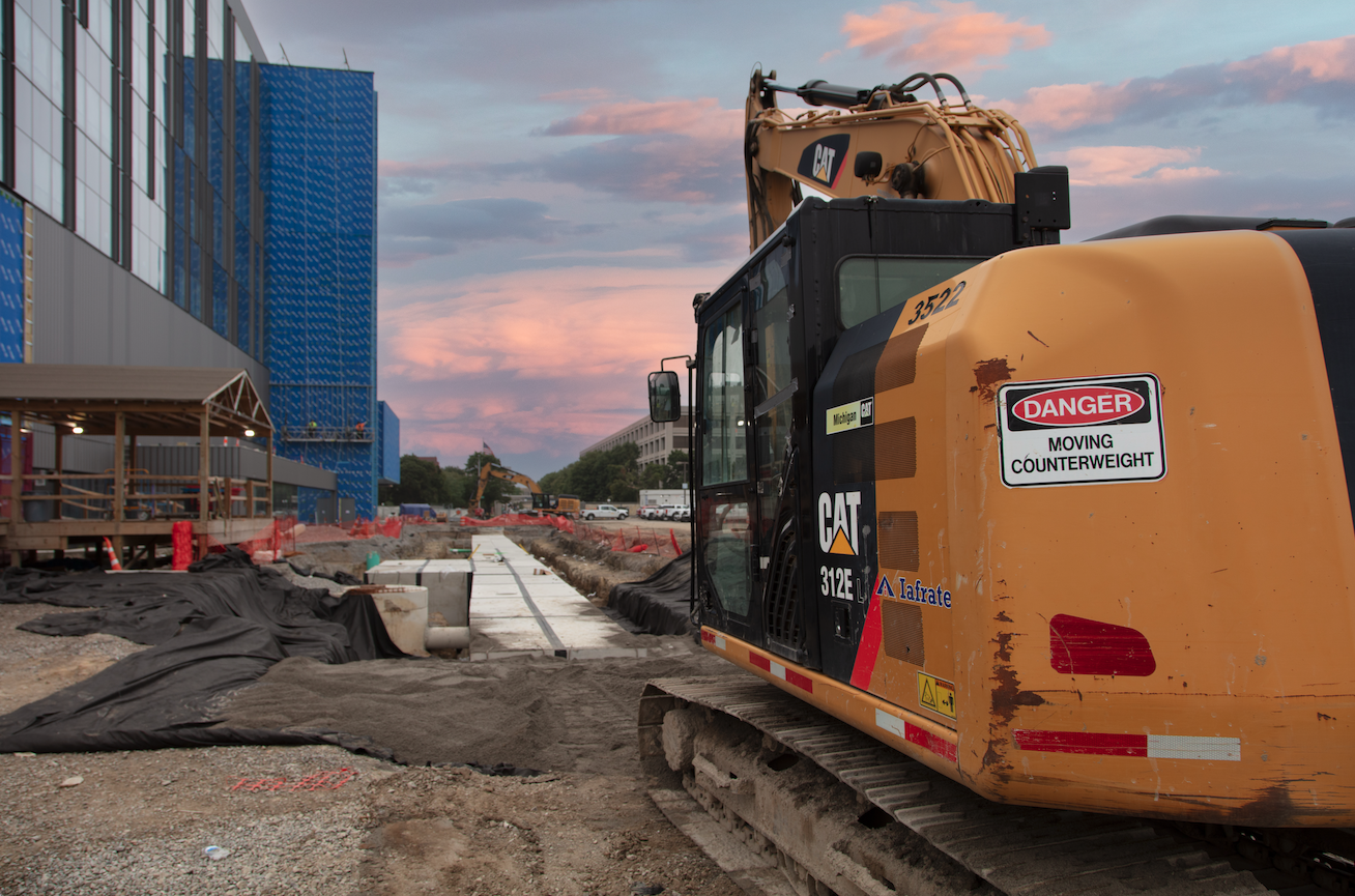 Construction site with a yellow Caterpillar excavator labeled '312E' in the foreground, and a partially built modern blue building with large glass windows in the background. The scene is during dusk with pink and purple sky colors.